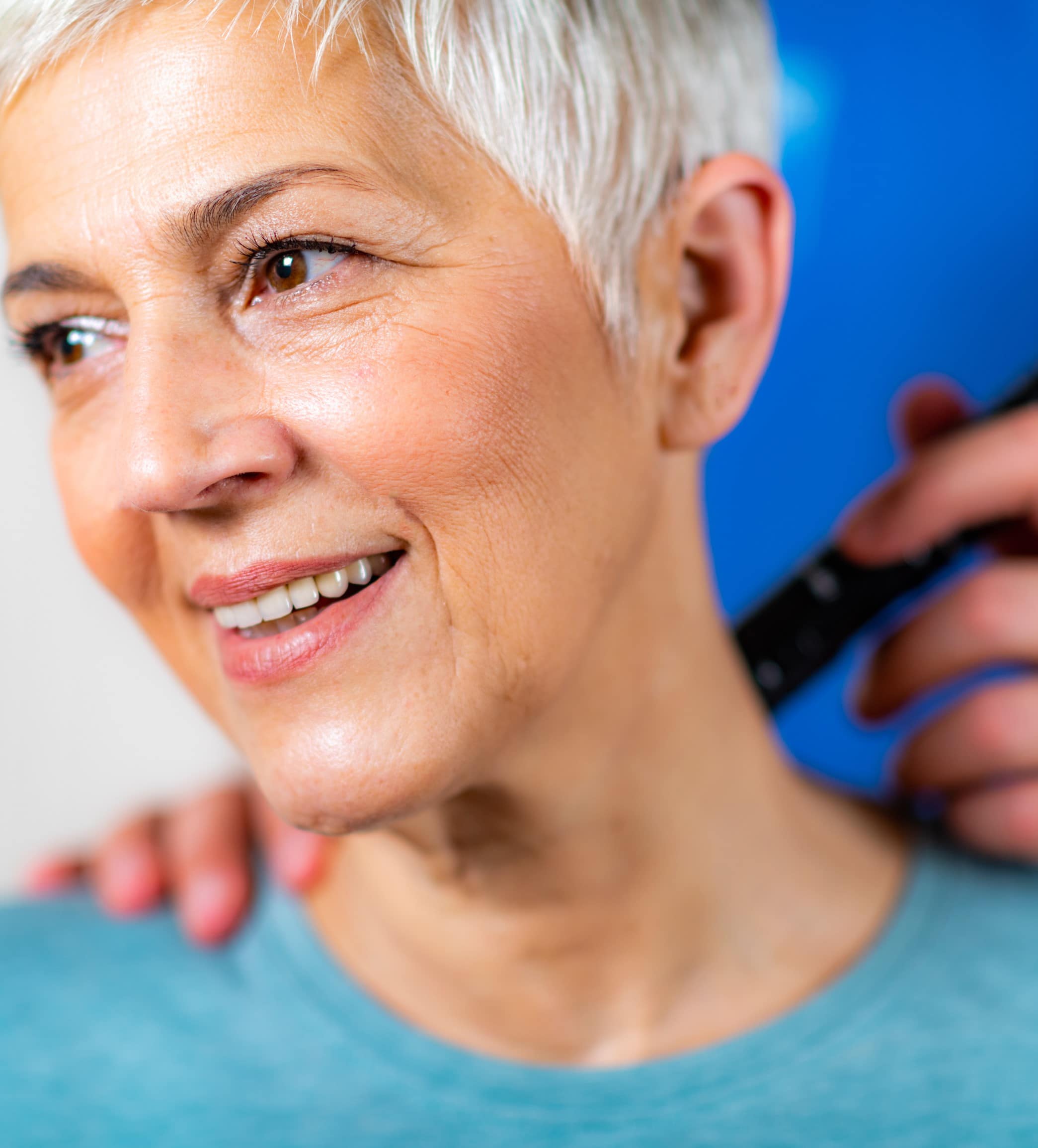 A smiling woman with short gray hair receiving shockwave treatment at the side of her neck and shoulder. She is seated upright in her regular clothing while a therapist's hand gently holds the small handheld treatment wand against her skin. She looks relaxed and comfortable; the treatment involves no needles and does not break the skin.